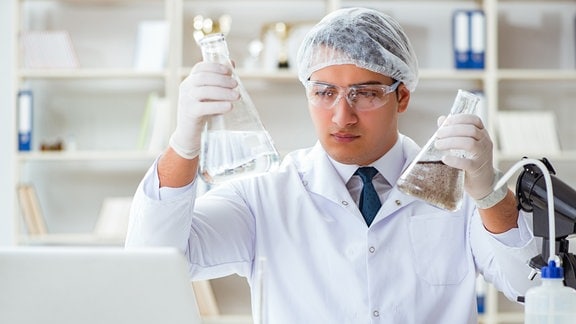 Junger Wissenschaftler mit Kittel, Haube und Schutzbrille hält zwei Glaskolben – einen mit klarem und einen mit bräunlichem Wasser – im Labor vor Laptop hoch und betrachtet Inhalt.