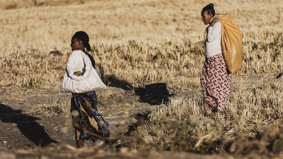 Zwei Frauen laufen mit Säcken auf dem Rücken auf einem sehr trockenen Feld