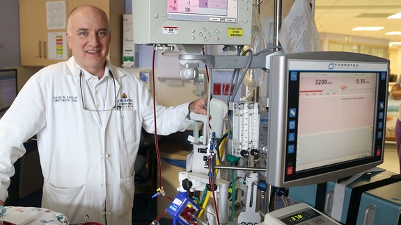Ein Arzt steht neben einer Herz-Lungen-Maschine und schaut lächelnd in die Kamera.