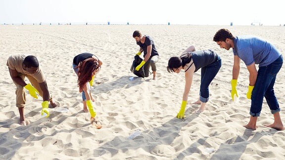 Junge Erwachsene sammeln Müll an einem Strand ein. Ansicht von vorn als Gruppe mit viel Sand bis zum Horizont des Bildes. Menschen in gebückter Haltung mit gelben, langen Gummihandschuhen.