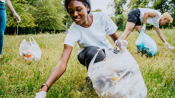 Gehockte Perspektive: Jung Frau sammelt freiwillig Müll ein im Park, gut gelaunt, im Hintegrund weitere Menschen