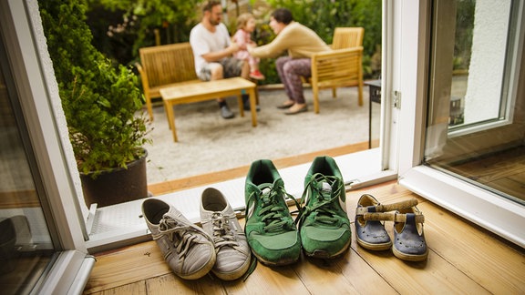 Gestellte Aufnahme: Die Schuhe einer jungen Familie stehen in einem Haus an der Terrassentuer, waehrend die Familie im Hintergrund auf einer Bank sitzt.