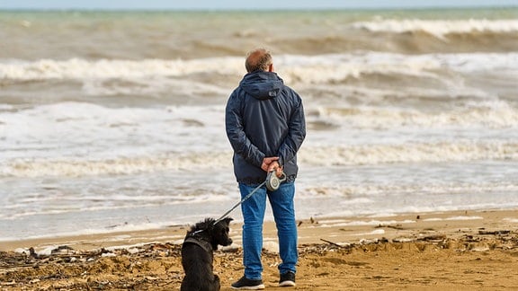 Alter Mann schaut auf das Meer hinaus, während er mit seinem Hund an der Leine durch den Sandstrand spazieren geht.