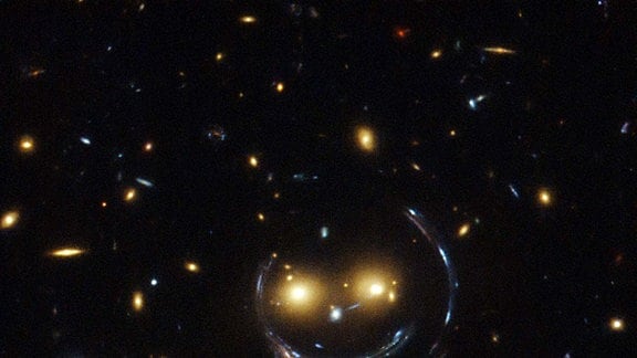 In der Mitte dieses Bildes, aufgenommen mit dem NASA/ESA Hubble Weltraumteleskop, befindet sich der Galaxienhaufen SDSS J1038+4849. In diesem speziellen Fall vom Gravitationslinsen-Effekt entsteht durch die Lichtbeugung ein Ring - der so genannte Einstein-Ring. Der Ring zeigt ein verzerrtes Bild einer Galaxie, die weit hinter der im Vordergrund befindlichen Galaxie liegt, die wie ein lächelndes Gesicht aussieht.