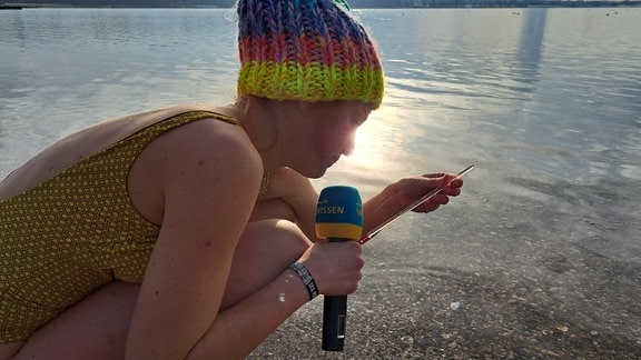 Eine Frau im gemusterten Badeanzug und mit bunter Wollmütze auf dem Kopf hält in der einen Hand ein Mikrofon und in der anderen ein Thermometer, mit dem sie die Wassertemperatur am Ufer eines Sees misst.