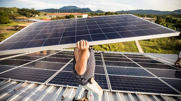 Ansicht von leicht oben in Weitwinkel-Perspektive: Mann trägt Photovoltaik-Panel auf einem Dach, Kopf von Panel verdeckt, im Hintergrund Häuser und Landschaft