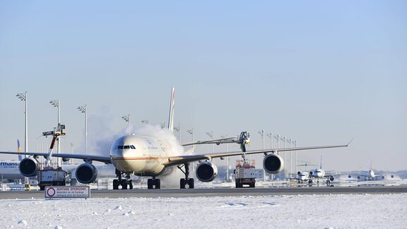 Flugzeug von vorn auf schneebedecktem Untergrund an Flughafen; Fahrzeuge sprühen nebelartige Substanz auf beide Seiten des Flugzeugs; blauer sonniger Himmel