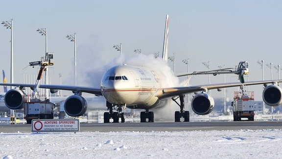 Flugzeug von vorn auf schneebedecktem Untergrund an Flughafen; Fahrzeuge sprühen nebelartige Substanz auf beide Seiten des Flugzeugs; blauer sonniger Himmel