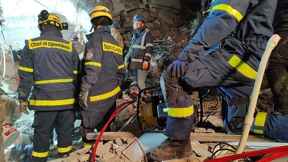 Vier Einsatzkräfte vom THW stehen inmitten der Trümmer eines bei einem Erdbeben eingestürzten Hauses.