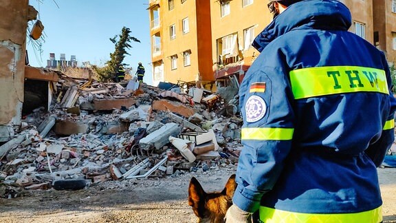 Eine Rettungskraft in THW-Kleidung steht mit einem Suchhund vor den Trümmern eines bei einem Erdbeben eingestürzten Hauses.