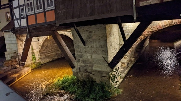 Krämerbrücke mit Fachwerkbebauung und tiefen Brückenbögen über Wasser. Die Bögen und das Wasser werden durch künstliches Licht erhellt.