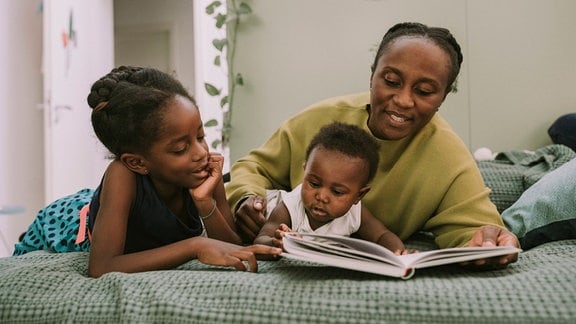 Mutter mit Kindern liegen im Bett und lesen ein Buch.