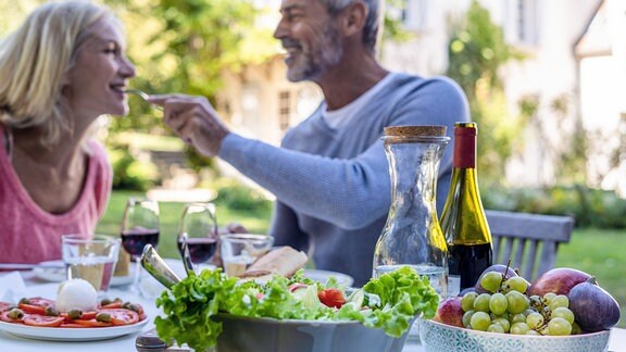 Lächelnder Mann mittleren Alters sitzt mit Frau mittleren Alters an einem Tisch im Freien und füttert Sie mit einer Gabel. Im Vordergrund auf dem Tisch sind Gemüse, Obst sowie Wein und Wasser angerichtet.