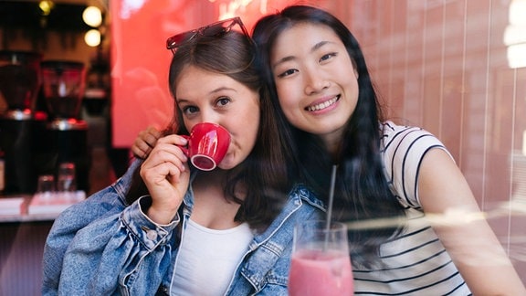 Zwei junge Frauen blicken angelehnt, freudig und ausgelassen in die Kamera, eine trinkt aus einem roten Tässchen Espresso
