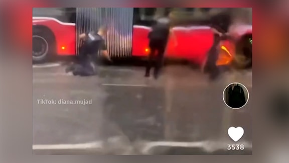 Unscharfer Screenshot von TikTok: Roter Gelenkbus auf Nasser Straße, davor Personen, knieender Polizist