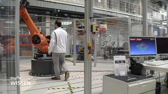 Ein Techniker geht zu einem Indutrieroboterarm. Rechts im Bild sind Computermonitore zu sehen.