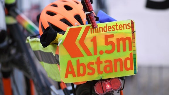 Nahaufnahme Fahrradkorb mit Helm auf Gepäckträger. An Korb leuchtgelbes Schild mit roter Schrift "mindestens 1,5 Meter Abstand" und Pfeil nach links. Hintergrund mit Vorderteil des Fahrrads unscharf.