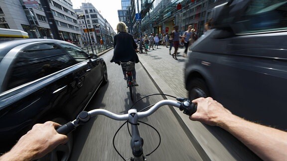 Ein Radfahrer wird im Stadtverkehr von einem nah vorbeifahrenden Auto ueberholt. Egoperspektive aus Radfahrer-Sicht mit armen und gebogenem Lenker im Vordergrund. Rechts ein parkendes Auto.
