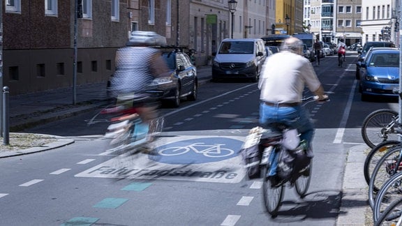 Zwei radfahrende Personen fahren mit dem Rücken zur Kamera in eine Fahrradstraße einer Großstadt. Bewegungsunschärfe. Parkende Autos am Straßenrand.