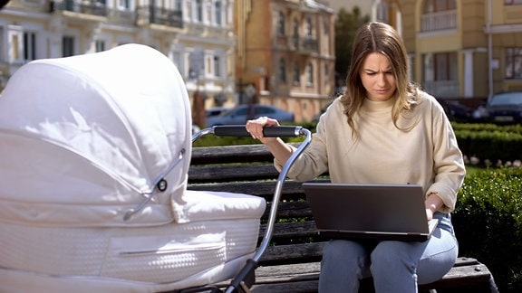 Junge Frau sitzt mit einer Hand an einem Kinderwagen auf einer Bank und schaut mit gerunzelter Stirn auf einen Laptop auf ihrem Schoß.