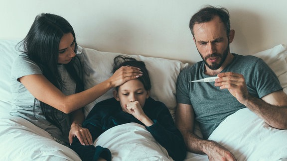 Familie mit Kind liegt krank im Bett, der Vater schaut auf ein Fieberthermometer, die Mutter hat eine Hand auf der Stirn des Kindes. Das Kind hustet in seine Faust.