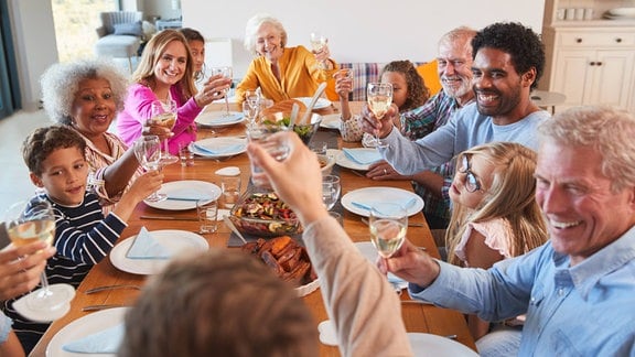Bunte erweiterte Familie sitzt um gedeckten Tisch und erhebt Gläser mit Wasser und Weißwein. Freudige Gesichter. Blick zur Person an Stirnkante, unscharf mittig im Vordergrund.