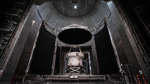 Das Orion-Raumschiff der amerikanischen Raumfahrtbehörde NASA in seiner Fertigungshalle. In der unteren Bildmitte befindet sich die weiße Raumkapsel. Ansonsten ist das Bild in betongrau gehalten.