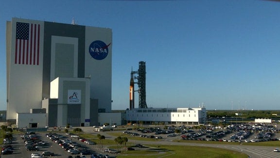 Die Mondrakete der NASA ist auf dem Kennedy Space Center in Florida unterwegs und verlässt das Vehicle Assembly Building für eine 4,2 Meilen lange Reise zum Startkomplex 39B. Alleine die Rakete ist ungefähr 98 Meter hoch, das Fahrzeug auf dem sie Transportiert wird ist ebenfalls circa sieben Meter hoch. Im Vergleich zur Fertigungshalle wirken sie aber winzig.