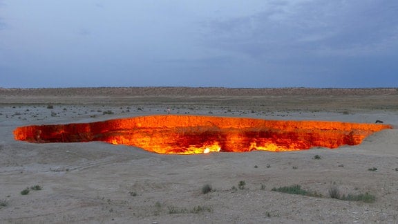 Wüstenlandschaft in der Dämmerung. Die obere Hälfte des Bildes wird von dem blaugrauen Himmel bestimmt, die untere von der Wüste mit einem großen Krater, in dem ein Feuer zu lodern scheint.
