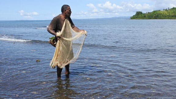 Seitenansicht: Mann mit Fischernetz steht im knöchelhohen Meereswasser, im Hintergrund grünes Ufer