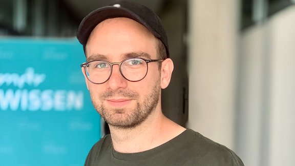 Mann mit bart, runder schwarzer Brille, schwarzem Pullover, schwarzem Basecap