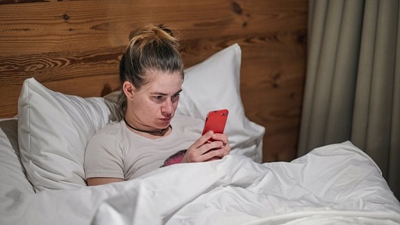 Junge Frau mit blonden, hochgesteckten Haaren liegt in Bett mit weißer Bettwäsche vor Holzwand und starrt auf rotes Smartphone, das sie mit beiden Händen festhält