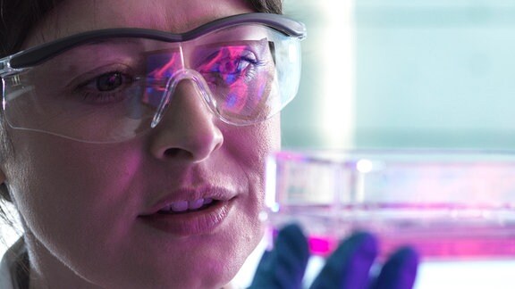 Eine Frau in einem Laborkontext trägt eine Schutzbrille und hält ein Schälchen in einer Hand.