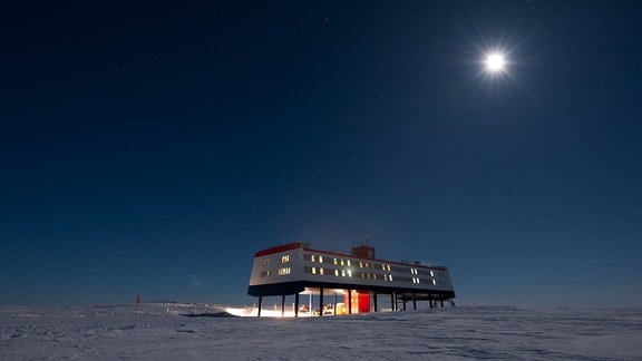 Die deutsche Antarktis-Forschungsstation Neumayer-Station III: Forschungsstation, die entfernt aussieht wie ein Schiff, auf Stelzen im Eis, Himmel dunkelblau-schwarz; in der Forschungsstation brennt Licht.