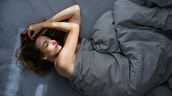 Draufsicht: Frau mit langen Haaren liegt mit nach oben gewinkelten Armen in dunkler Bettwäsche verschlungen im Bett, Augen geschlossen.