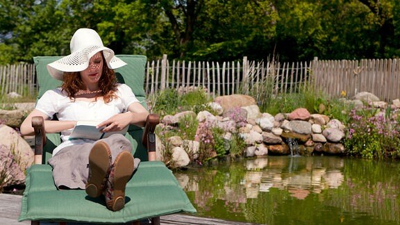 Eine junge Frau mit Sonnenhut liest auf einem Liegestuhl ein Buch, neben ihr ein Teich