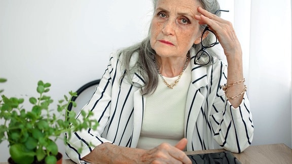 Müde ältere grauhaarige Geschäftsfrau in gestreifter Jacke mit Brille arbeitet in ihrem Büro, sitzt am Schreibtisch und fühlt sich aufgrund der Wechseljahre schlecht.