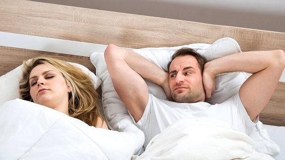 Ein Mann hält sich die Ohren zu während eine Frau neben ihm schläft