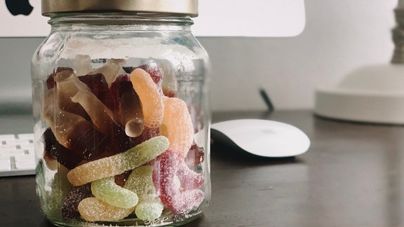 Ein Glas mit Schraubdeckel mit Fruchtgummi – Colafläschchen und bunte gezuckerte Würmer – steht auf einem dunklen Schreibtisch. Im Hintergrund unscharf teilweise ein Computer mit Maus, Tastatur und Fuß einer Schreibtischlampe.