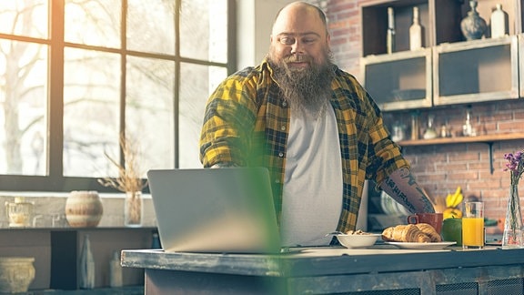 Fröhlicher Mann mit langem Bart und größerem Bauch steht an Laptop am Küchentisch, daneben Croissant, Orangensaft, Müsli. Sonnige Stimmung.