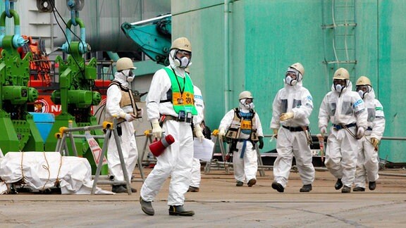 Arbeitende auf dem Gelände eines Atomkraftwerkes in weißen Schutzanzügen, Schutzhelmen und Atemschutzmasken.