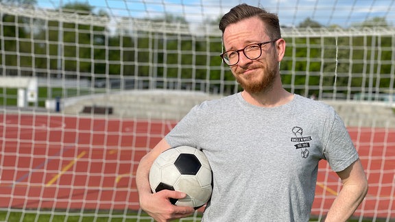 Bärtiger Mann mit Brille in grauem T-Shirt vor einem Fußballtor