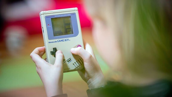 Kind spielt auf Gameboy der allerersten Generation das Spiel Tetis. Hintergrund farbig unscharf, Vordergrund unscharfer Kopf des Kinds, Blick über Schulter.