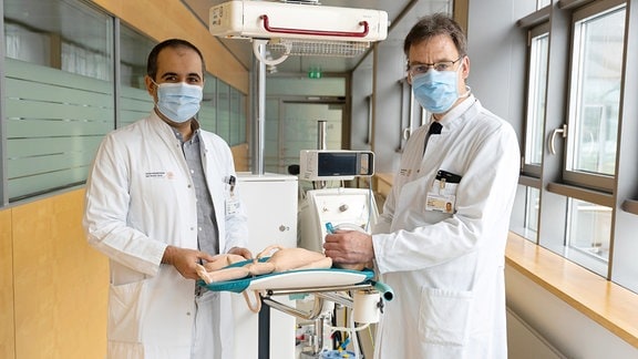 Zwei Männer in Ärztekitteln stehen in einem Gang in einem Krankenhaus vor einem Tisch mit einer Plastikpuppe darauf.  