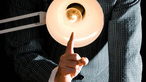 Verhörsituation: Lampe auf Betrachter gerichtet, dahinter Mann im Anzug, erhobener Zeigefinger vor Lampe.
