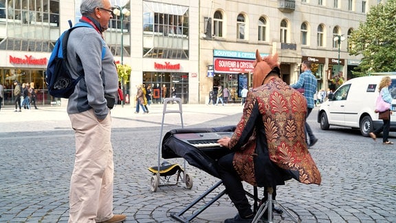 Straßenmusiker mit Perdekopf-Maske spielt E-Piano in Prager Fußgängerzone. Mann mit grauen Haaren, Brille, Rucksack und Händen in den Taschen steht interessiert daneben.