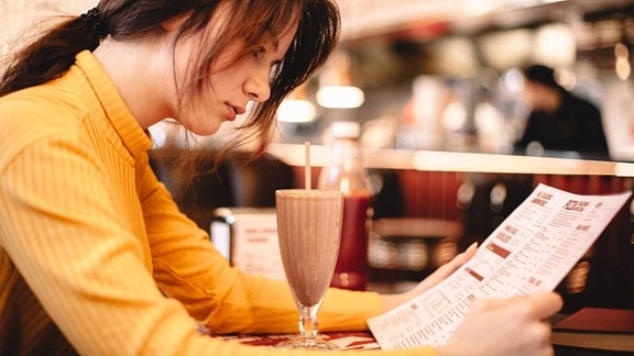 Seitenansicht: Junge Frau mit Pferdeschwanz sitzt in Café vor Milchshake und studiert eine Speisekarte, Hintergrund unscharf