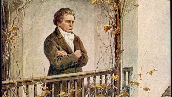 Ludwig van Beethoven seitlich: Mann mit lockigen Haaren steht mit verschränkten Armen auf einem Balkon von Haus mit herbstlichem Laub umgeben.