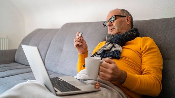 Mann sitzt auf der Couch mit Decke und Laptop auf dem Schoß und hält eine Thermometer und eine Teetasse in den Händen.