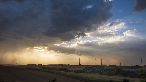 Windräder auf dem hügeligen Land, darüber eine Gewitterwolke, orangefarbenes Gegenlicht der Sonne, teilweise blauer Himmel